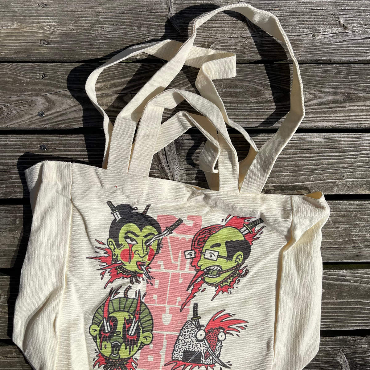 „Namakubi“ Tote Bag von Heldbergs Games mit vier ikonischen Köpfen, aus 16-Unzen-Baumwolle. Maße 32 x 33 x 16 cm, mit Magnetverschluss und Innentasche. Japanisches Design trifft auf praktischen Stil.